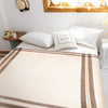 Bed with a neatly arranged cream and brown-striped blanket, white pillows including one with FEEL GOOD text, in a cozy room with a wooden window, hat, phone, and nightstand.