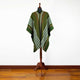 A green poncho with white and orange stripes hangs on a wooden dress form. The setting includes a wooden floor and a plain white background.
