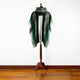 A green and grey cloak with decorative patterns displayed on a wooden stand, situated on a polished wooden floor against a plain white wall.
