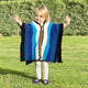 Wuachapa llama wool hooded poncho – a child, wearing a multicolored striped poncho, stands with arms outstretched on a grassy lawn with hedges in the background