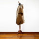A brown woven poncho with white geometric patterns displayed on a wooden mannequin stand, set in a room with wooden flooring against a plain white wall.