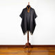 A black poncho with tassel trim is displayed on a wooden stand, featuring a colorful embroidered neckline, in a room with wooden flooring and a plain white background.