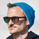 A person wears sunglasses labeled HAWKERS and a blue beanie, standing outdoors on a sunny day. Their expression is neutral, and they appear relaxed in a casual setting.