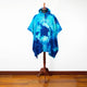 A blue tie-dye poncho with a hood is displayed on a wooden mannequin stand, situated in a room with polished wooden flooring and a plain white wall background.