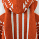 A soft knit sweater with a hood, featuring bold orange and white patterns, displays vertical stripes and geometric designs, creating a cozy, autumnal feel.