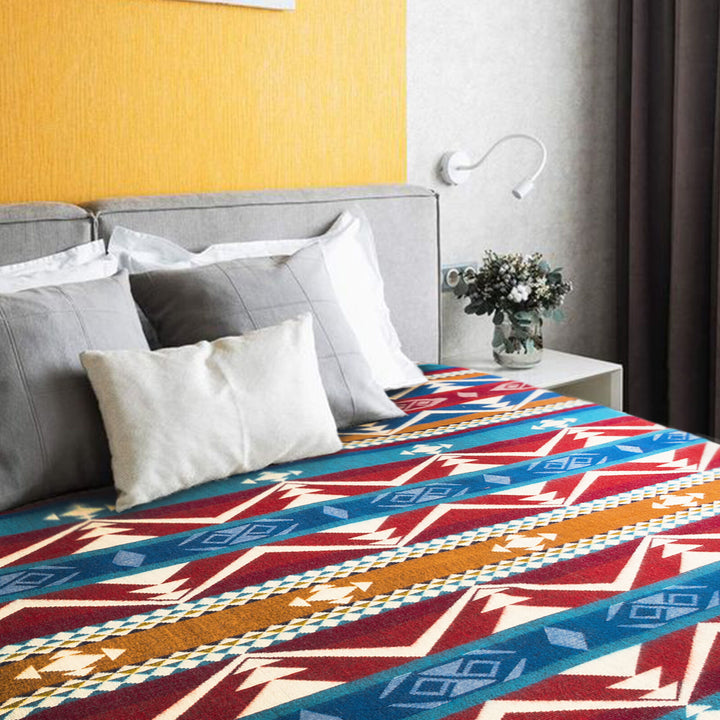 A colorful patterned blanket covers a bed adorned with gray and white pillows. A bedside table holds a vase with flowers, set against a yellow and gray wall backdrop.