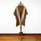 A brown poncho with a hood and geometric white patterns hangs on a wooden stand in a room with a wooden floor and white wall backdrop.