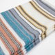Nongora baby alpaca wool blanket – a folded, multi-colored striped fabric with hues of teal, orange, gray, and yellow is laid on a plain white surface. The vibrant lines create a harmonious pattern