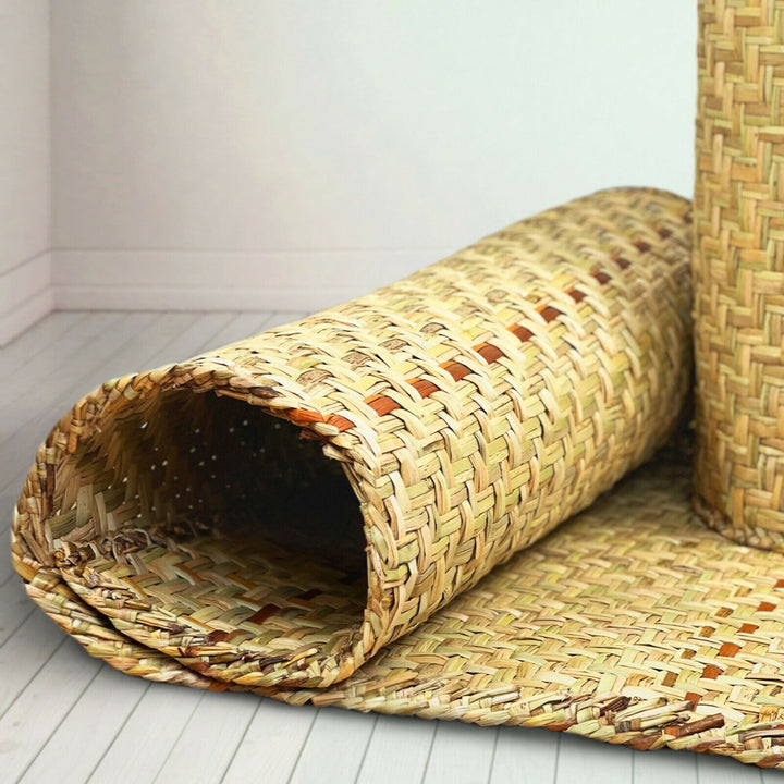 A rolled woven straw mat lies partially unrolled on a light-colored wooden floor, featuring a detailed pattern of interlaced natural fibers in a minimalist room setting.