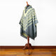A decorative poncho with geometric patterns is displayed on a wooden mannequin stand, set against a plain white wall on a hardwood floor.