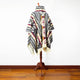 A multicolored poncho with intricate geometric patterns and fringed edges is displayed on a wooden stand against a white background and wooden floor.