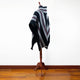 A striped poncho is draped over a wooden mannequin stand. The poncho features black, white, and red diagonal patterns. The setting includes a wooden floor and a plain white wall.