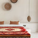 A bed covered with a red and white geometric-patterned blanket is set in a minimalist room with basket wall decor and a woven hanging light.