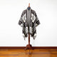 A black and white patterned poncho rests on a wooden mannequin stand, displayed in a room with wooden flooring and a plain white wall background.