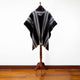 A black and gray striped poncho is displayed on a wooden mannequin stand with a white background and hardwood floor.