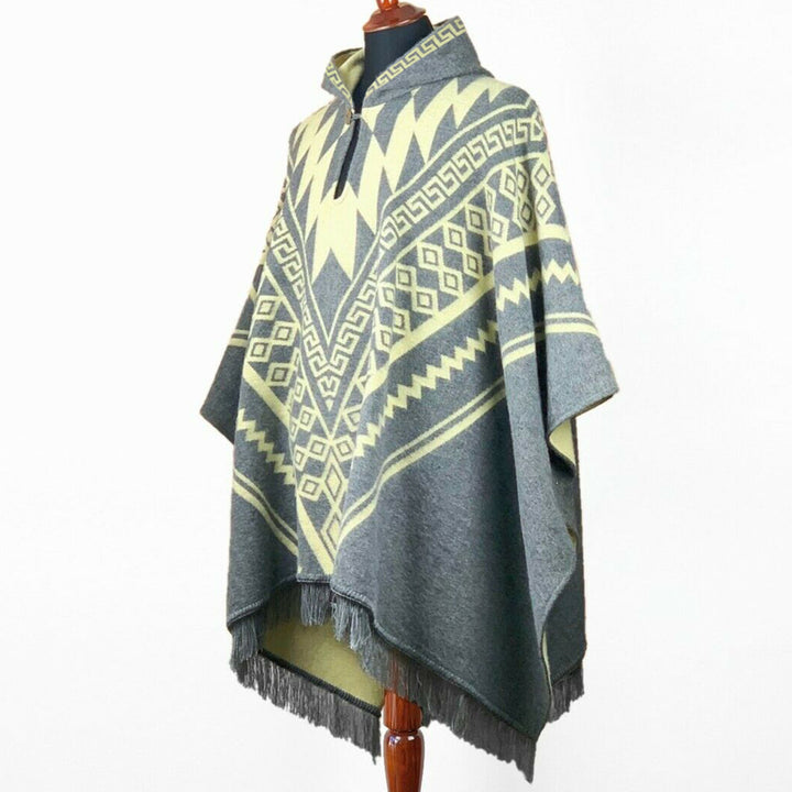 A poncho with geometric patterns is displayed on a mannequin. The poncho features a zigzag and diamond design in gray and cream, with fringed edges.
