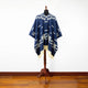 A dark blue poncho with white geometric patterns is displayed on a wooden mannequin stand. The garment's fringed edges drape elegantly, set against a white wall and wooden floor.