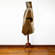 A brown wool poncho with geometric patterns drapes over a mannequin on a wooden stand, set on a wooden floor against a plain white wall.