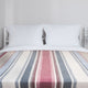 Pindo baby alpaca wool blanket – bed with white pillows and a striped blanket featuring red, blue, cream, and beige colors; set against a plain light-colored wall in a minimalistic bedroom