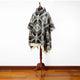 A poncho with geometric patterns and fringe edges is displayed on a wooden mannequin stand, set against a simple white wall and wooden floor.