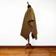 A woven poncho, featuring diagonal brown and yellow stripes, is draped over a mannequin stand. The setting includes a wooden floor against a plain white wall backdrop.