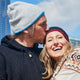 A man kisses a woman’s forehead as she smiles warmly. They are outdoors, dressed in winter clothes with a clear, blue sky and buildings in the background.