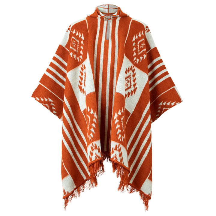 A vibrant poncho displays orange and white geometric patterns, featuring stripes and diamond shapes. It has fringed edges and a high collar with a decorative clasp, set against a plain background.