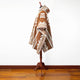A brown and beige patterned poncho drapes on a wooden mannequin stand against a white wall, positioned on a polished hardwood floor.