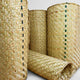 Woven mats, rolled standing upright, display intricate straw patterns with occasional green and black threads. They rest on a plain surface, emphasizing their detailed texture and craftsmanship.