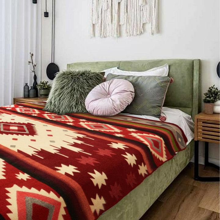 A bed is adorned with red and beige geometric-patterned blanket, surrounded by green, pink, and gray pillows. A bedside table with plants and modern decor enhances the cozy bedroom setting.