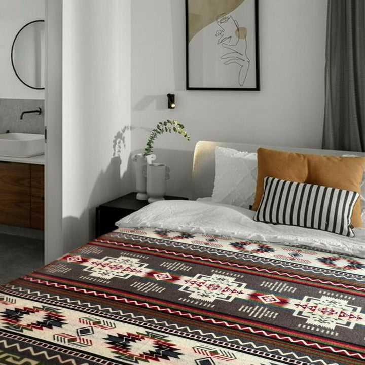 Bed adorned with a patterned blanket and cushions; situated in a modern bedroom with neutral decor. A round mirror and sink are visible in the adjacent bathroom.