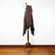 A brown wool cloak with fringe hangs on a wooden mannequin stand in a room with wooden flooring and white walls.
