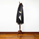 A dark gray wool poncho with a hood and fringe edges is displayed on a wooden stand, set in a room with a white wall and wooden floor.