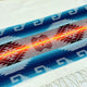A woven textile displays a vibrant geometric pattern with blue, orange, and red hues, bordered by white fringe. The intricate design features zigzags and diamond shapes.