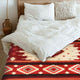 A bed is adorned with white pillows and sheets, featuring a red and beige geometric patterned blanket. An earthy vase is placed on the wooden floor beside it.