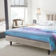 Tinizaray baby alpaca wool blanket – bed is made with colorful blanket. Lamp and photo frame sit on nightstand. Wooden chair by window with soft light creates a cozy bedroom atmosphere