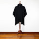 Achema llama wool hooded poncho – a black poncho drapes over a wooden mannequin stand, displayed in a minimalist setting with a white wall and wooden floor