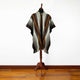 A patterned poncho with geometric designs hangs on a wooden mannequin stand in a room with a wooden floor, against a plain white wall.