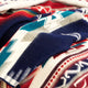 A textured blanket displays geometric patterns in blue, red, and white hues, draped naturally. The intricate zigzag and stepped designs create a visually dynamic, cozy appearance in an indoor setting.