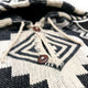Hooded garment with black and beige geometric patterns features two brown buttons and a drawstring. The close-up view emphasizes its woven texture and intricate design elements.