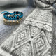 Two woven bracelets, one blue with black beads and one beige with round stones, rest on a soft, gray patterned fabric, displaying intricate geometric designs.