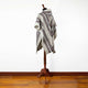 A woolen poncho with diagonal stripes in gray, black, and red is displayed on a wooden mannequin stand, set against a plain white wall and hardwood floor.
