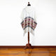 A white hooded poncho with colorful geometric patterns drapes over a wooden mannequin stand, set against a plain white wall and wooden floor.