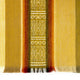 A woven textile displays horizontal bands of yellow, orange, and brown with intricate central diamond patterns. The fabric features fringed edges, providing a traditional, handcrafted appearance.