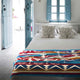 A bed with a colorful geometric-patterned blanket and decorative pillows featuring tree designs is positioned in a bright room. Blue doors and windows enhance the airy, coastal ambiance.