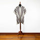 A gray poncho with red and black stripes hangs on a wooden mannequin stand. The setting is a room with a white wall and light wood flooring.