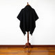 A black poncho drapes on a wooden mannequin stand, set against a white wall and a polished wood floor.