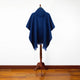 A blue poncho with fringed edges is draped on a wooden stand. It has a hood and is displayed against a plain white wall on a wooden floor.