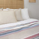 Pindo baby alpaca wool blanket – a neatly made bed is adorned with a textured, natural-colored headboard and layered with pillows. A striped blanket in muted red, blue, and beige accents the white bedding