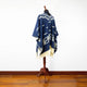 A blue poncho with white geometric patterns is displayed on a wooden mannequin. The poncho has fringed edges and is set against a plain white wall, on a wooden floor.